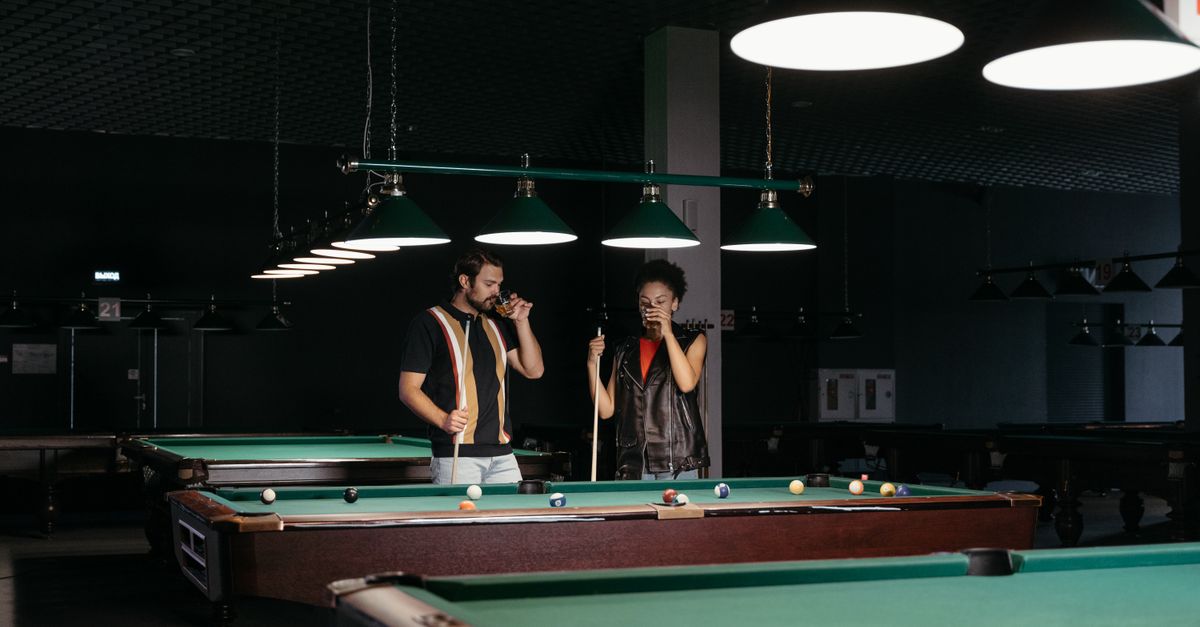 A man and woman playing billiards, enjoying drinks in a cozy indoor setting.